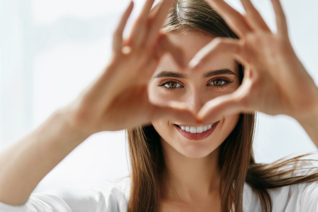 A beautiful young woman with healthy skin making a heart sign in front of her bright glowing eyes.