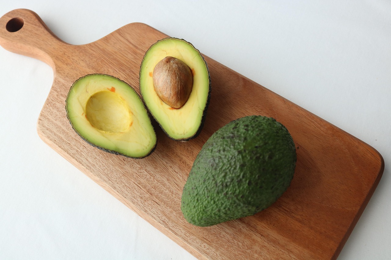 A sliced open healthy avocado sitting on a chopping board next to a fresh unopened avocado.